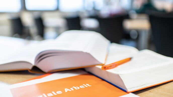 Foto von Büchern, die aufgeschlagen übereinander auf dem Tisch liegen. Davor ein Heft mit der Aufschrift "Soziale Arbeit".