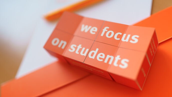 Foto eines aufgeklappten Klappwürfels mit dem Aufdruck "we focus on students", der auf Unterlagen liegt. __ Opened folding cube with the imprint "we focus on students" lies on documents.