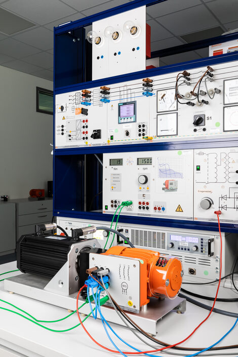 Foto eines Prüfstands für Elektromotoren mit Spannungsversorgung und Messgeräten, davor ein orangefarbener Drehstrommotor mit gekuppelter Lastmaschine auf dem Tisch.