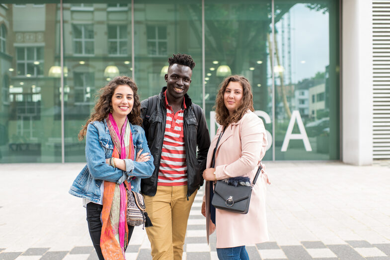 Foto von zwei Studentinnen und einem Studenten, die nebeneinander vor dem Mensagebäude stehen. Sie lachen in die Kamera.__Two female and one male students stand side by side in front of the canteen building. They laugh at the camera.