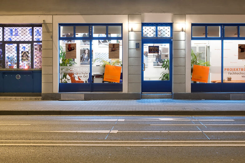 Foto des Projektbüros der Hochschule vor Ort von außen: Eine Hauswand mit großen blau gerahmten Fenstern, durch die man ins Gebäude sieht