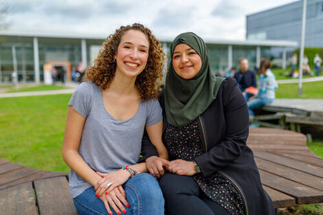 Foto von zwei Studentinnen, die nah beieinander auf Holzbänken auf dem Campus sitzen und in die Kamera lachen.__Two female students sit close together on wooden benches on the campus and smiling at the camera.