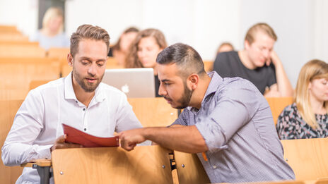 Photo of students in the lecture hall, in the foreground a student shows another student something in a folder. Other students are sitting in the background.
