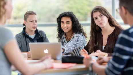 Photo of five students at a group table. The focus is on a female student and a male student sitting next to each other and looking at a laptop together.