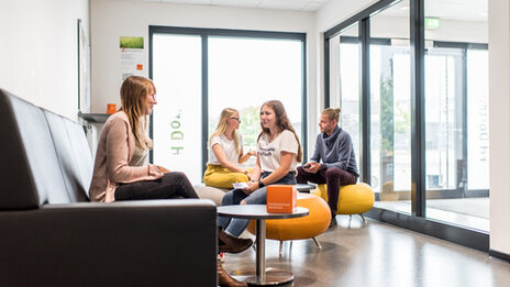 Foto eines Warteraum, in dem mehrere Personen sitzen und sich unterhalten.__Young people sittin in a waiting room and talking.