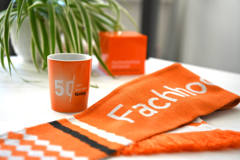 Eine Tasse und ein Schal liegen auf einem Tisch. Beide sind orange. Auf dem Schal steht "Fachhochschule" und auf der Tasse "Wir werden fünzig".