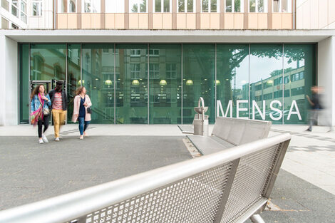 Drei Studierende kommen aus der neuen Mensa an der Sonnenstraße. Groß im Bild ist die Glasfassade der Mensa.