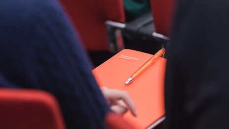 Foto einer Person auf einem Stuhl bei einer Veranstaltung mit FH-Unterlagen auf dem Schoß. __ Person sits on a chair at an event with FH documents on their lap.