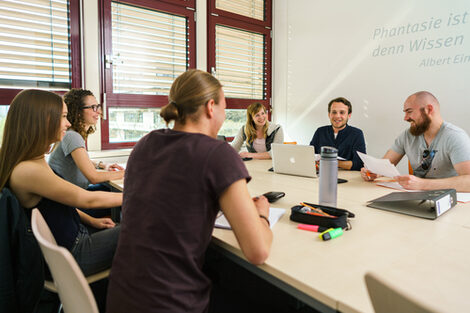 Photo of six students at a group table exchanging ideas. Working materials are on the table __Six students at a group table exchange ideas. Working materials are on the table.