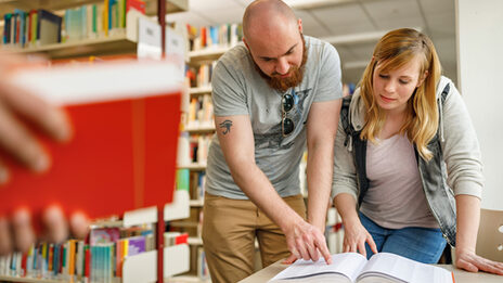 Foto einer Studentin und eines Studenten in der Bibliothek. Er zeigt ihr etwas in einem aufgeschlagenen Buch.__In the library, a student shows something to another student in an open book.