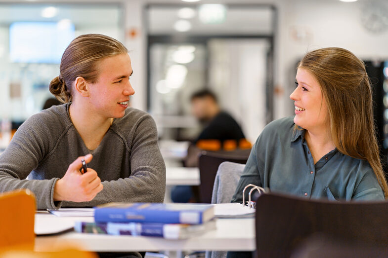 Photo of a female student and a male student sitting next to each other at a table, they laugh at each other. Books are on the table.__Student sit next to each other at a table, they laugh at each other. Books are on the table.<br><br>