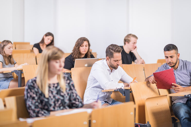 Foto von Sitzreihen im Hörsaal mit mehreren Studierenden. Alle schauen konzentriert auf ihre Tische und schreiben, lesen oder tippen am Laptop.