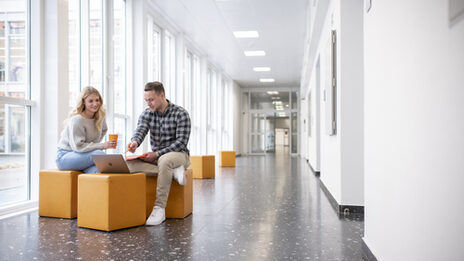 Photo of a female student and a male student. They are sitting in the hallway at the FH and talking. The student is pointing at something on his laptop.