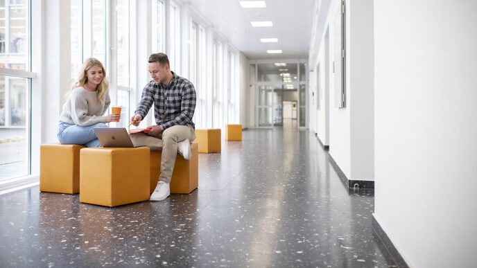 Photo of a female student and a male student. They are sitting in the hallway at the FH and talking. The student is pointing at something on his laptop.