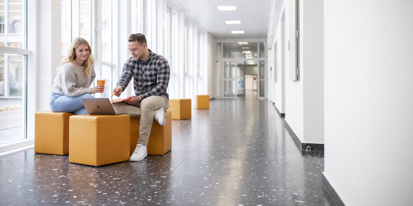 Foto von einer Studentin und einem Studenten. Sie sitzen im Flur in der FH und unterhalten sich. Der Student zeigt auf etwas am Laptop.