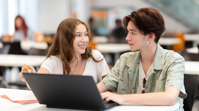 Foto von einer Schülerin und einem Schüler, die nebeneinander an einem Tisch sitzen und sich anschauen. Vor ihnen steht ein Laptop.