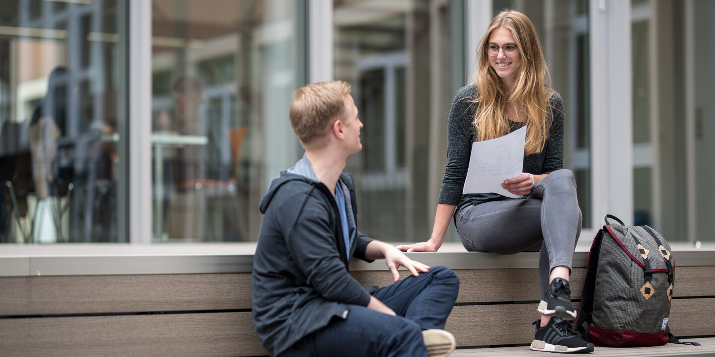 Foto einer Studentin und eines Studenten, die auf dem Campus auf Holzstufen sitzen und sich unterhalten. Die Studentin hält einen Zettel in der Hand.__One male and one female student sitting on wooden steps and talking, the female student is holding a note.