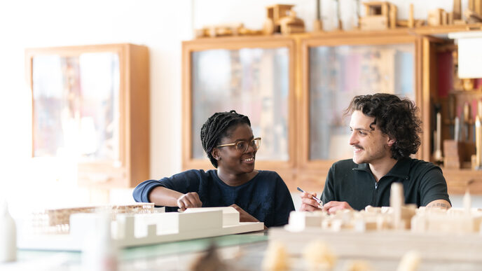 Foto von zwei Studierenden, sie sitzen an einem Tisch in der Holzwerkstatt und unterhalten sich, während sie an Modellen aus Holz arbeiten
