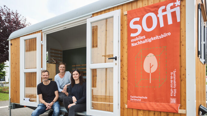 Man sieht 3 Personen, die in einem Bauwagen sitzen, welchen man über eine kleine Treppe betreten. An dem Bauwagen ist ein großes Plakat befestigt auf dem „Soffi mobiles Nachhaltigkeitsbüro“ steht.