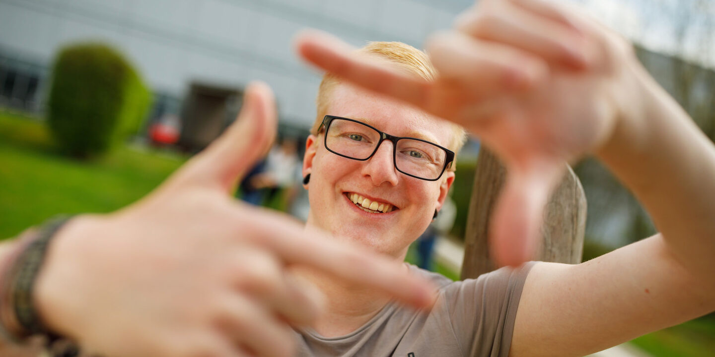 Foto eines Studenten, der mit Zeigefingern und Daumen vor sich ein Quadrat formt und dadurch guckt.