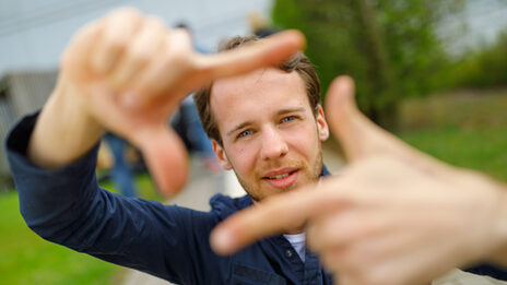 Foto eines Studenten, der mit Zeigefingern und Daumen vor sich ein Quadrat formt und dadurch guckt.