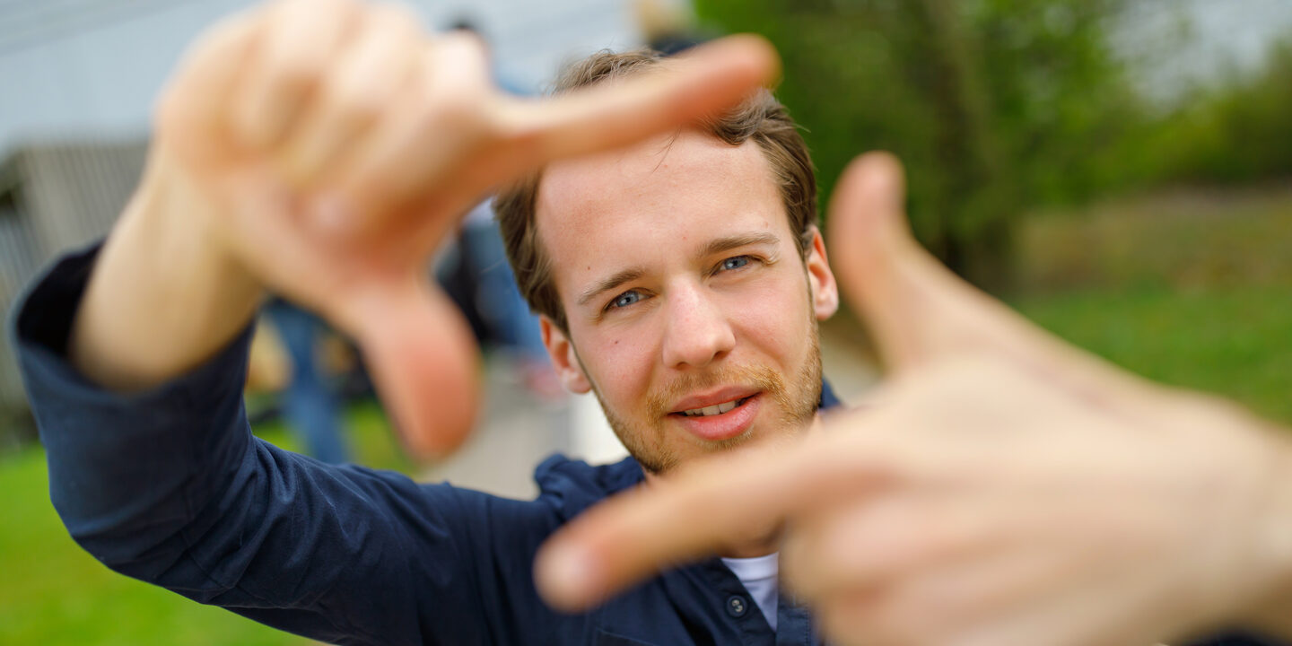 Foto eines Studenten, der mit Zeigefingern und Daumen vor sich ein Quadrat formt und dadurch guckt.
