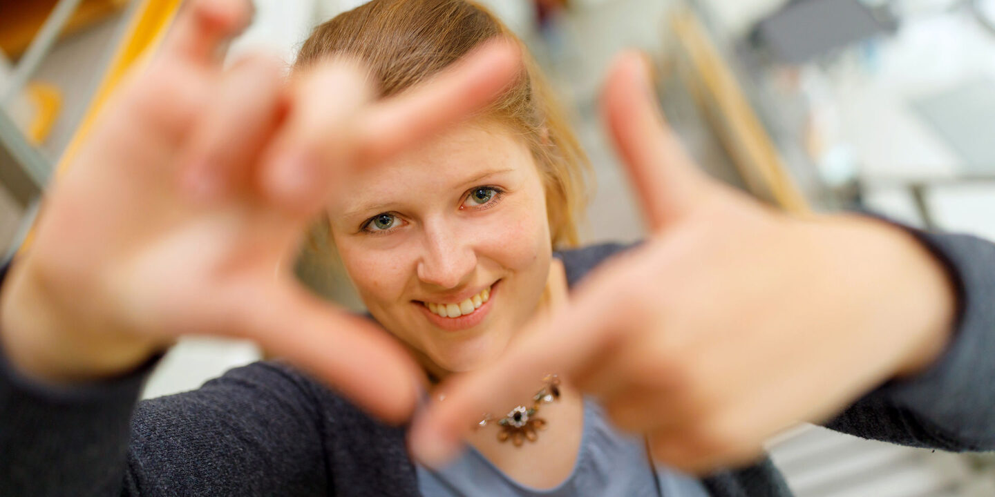 Foto einer Studentin, die mit Zeigefingern und Daumen vor sich ein Quadrat formt und dadurch guckt.