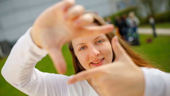 Foto einer Studentin, die mit Zeigefingern und Daumen vor sich ein Quadrat formt und dadurch guckt.