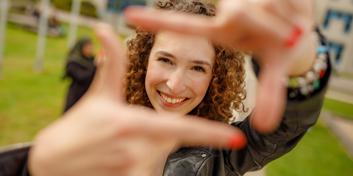 Foto einer Studentin, die mit Zeigefingern und Daumen vor sich ein Quadrat formt und dadurch guckt.