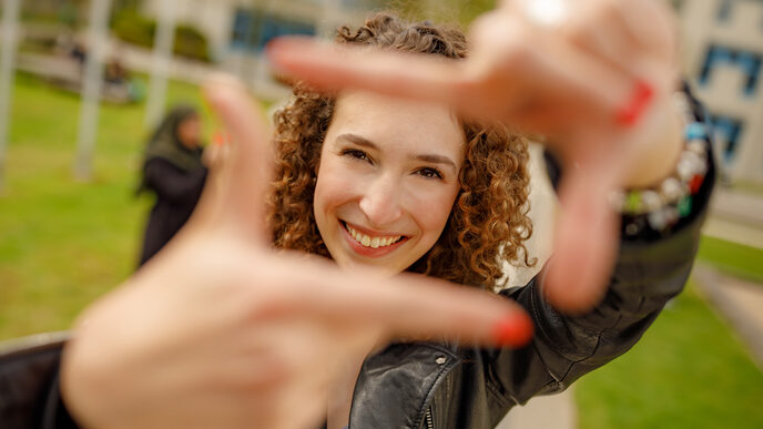 Foto einer Studentin, die mit Zeigefingern und Daumen vor sich ein Quadrat formt und dadurch guckt.