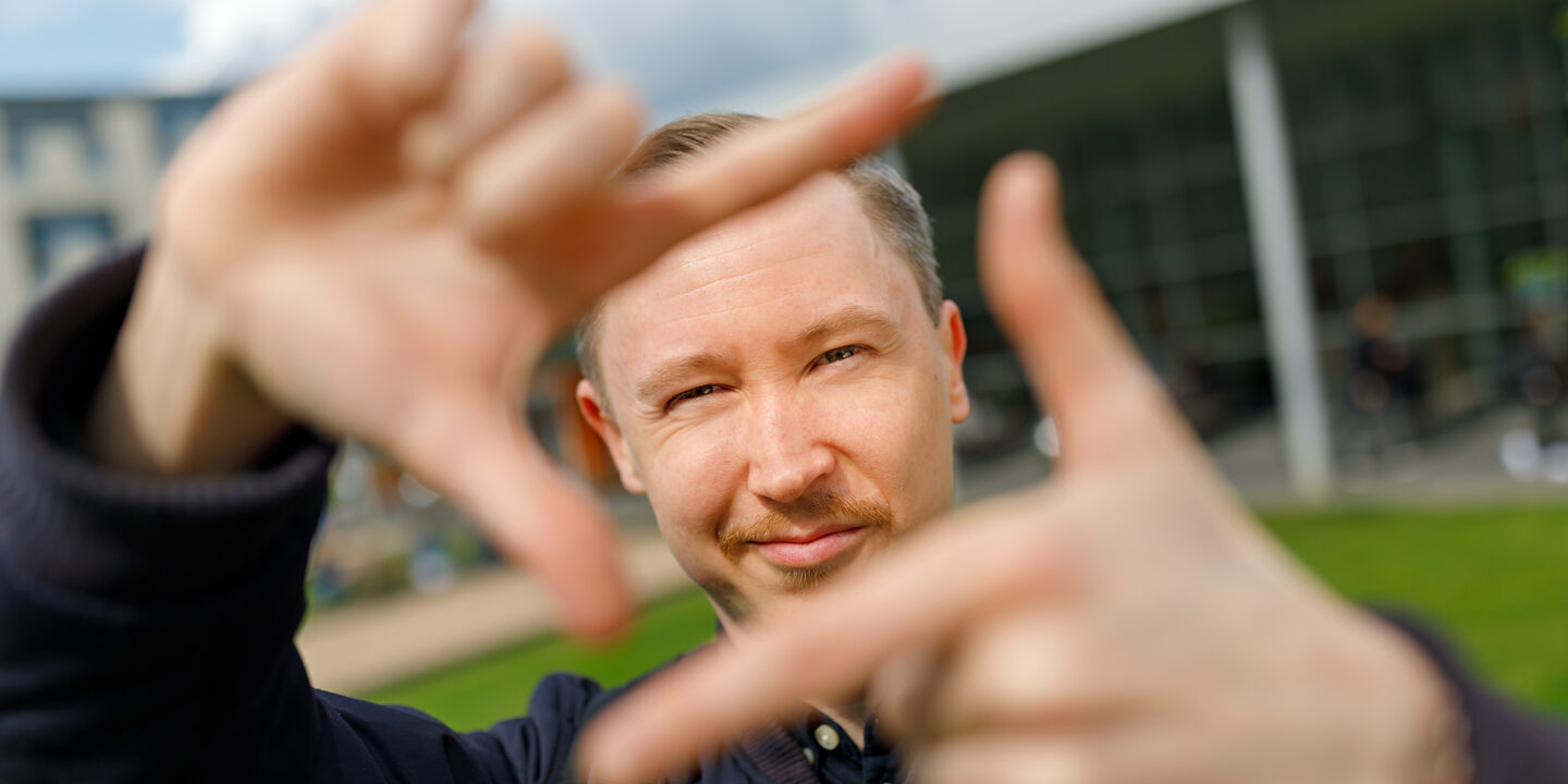 Foto eines Studenten, der mit Zeigefingern und Daumen vor sich ein Quadrat formt und dadurch guckt.
