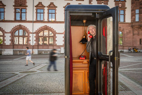 Auf dem Platz vor einem Gebäude steht eine schwarze Telefonzelle, darin zu sehen ist eine Person, die den Hörer eine roten Telefons ans Ohr hält. Das Telefon steht auf einer Holzkommode, darüber hängt eine Lampe.