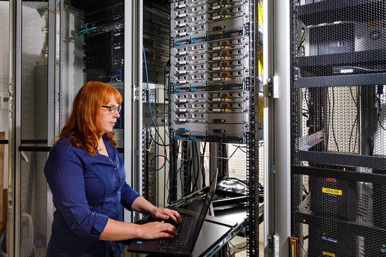 Foto einer Auszubildende vor einem großen Schrank mit Servern, sie bedient einen Laptop. __ <br>A trainee stands in front of a large cabinet with servers and operates a laptop.