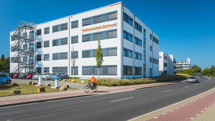 Photo of a Fachhochschule Dortmund building at Emil-Figge-Straße 38b with a view of the illuminated FH logo and the street and parking lot in the foreground.