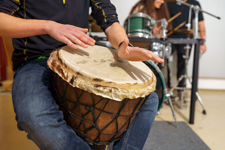 Foto eines Mannes, der im Sitzen auf einer Djembe-Trommel spielt, sein Kopf ist nicht mit auf dem Bild. Im Hintergrund spielen zwei Personen am Schlagzeug.