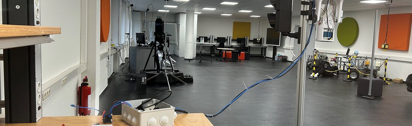 Ein Raum mit einem Holztisch im Vordergrund. Auf dem Tisch stehen verschiedene elektronische Geräte, darunter eine Ampel auf einem Ständer. Im Hintergrund sind weitere technische Geräte und Computerbildschirme zu erkennen. Der Raum ist ein Labor oder ein Forschungsraum.