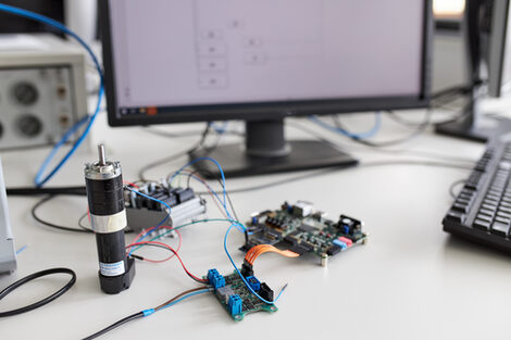 Bei diesem Arbeitsplatz wird ein kleiner Motor angesteuert und geregelt. Im Hintergrund steht ein eingeschalteter PC-Bildschirm. Auf dem Bildschirm wird ein Schaltplan angezeigt.