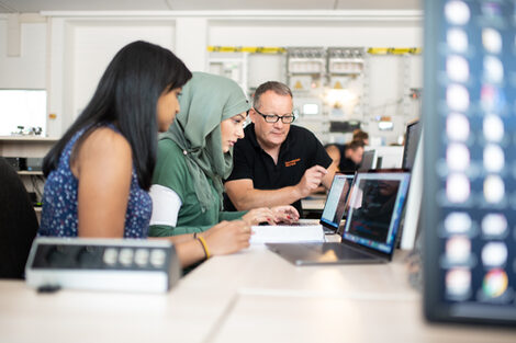 Foto von zwei Studentinnen und einem Professor im Labor für Energieautomation und Netzführung. Sie sitzen nebeneinander und schauen gemeinsam auf den Bildschirm eines Laptops.