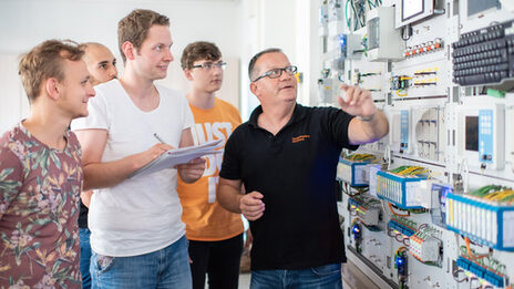 Foto eines Professors mit seinen Studierenden. Sie stehen vor einer Versuchswand der Elektrotechnik, der Professor zeigt auf ein Gerät. Einer der Studierenden macht sich Notizen.