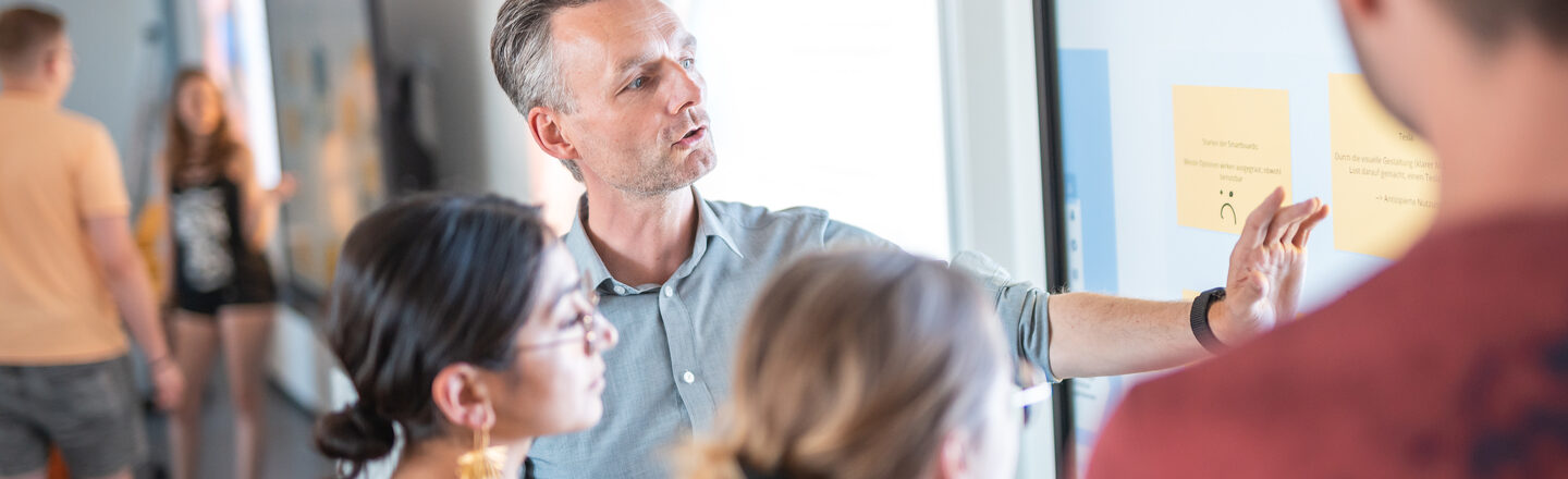 Photo of a group of students standing next to their professor at the whiteboard, who is explaining something to them. In the background, two other students are standing together at a board