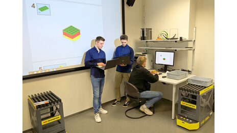 Two people are standing in front of a screen with laptops. A third person is sitting in front of a PC at a workstation.