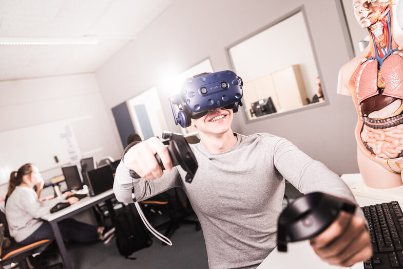 Foto von einem Studenten, der eine VR-Brille auf hat und Controller in der Hand hält, er sitzt an einem Rechnerarbeitsplatz. __A student has VR glasses on and controller in his hands, is sitting at a computer workstation.