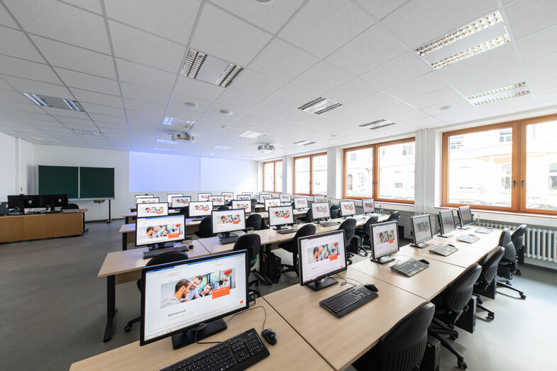 Ein Foto von einem Computerraum. Man sieht mehrere Tischreihen hintereinanderstehen, auf jedem Tisch steht jeweils ein Computer. Im Hintergrund sieht man eine Tafel und einen Beamer, der etwas an die Wand projiziert.