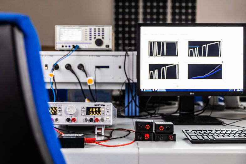 Photo of a test setup. On a table to the right is a screen with measurement results, to the left are wired measuring devices.