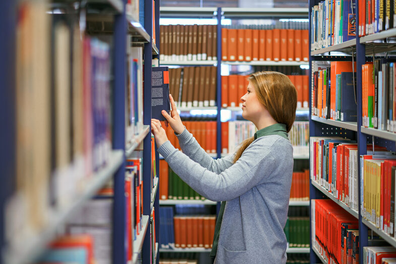 Foto einer Studentin in einer Bücherreihe der Bibliothek. Sie ist zum linken Regal gewandt und holt ein Buch raus.