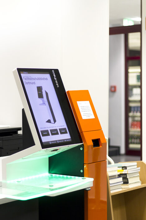 Foto eines Selbstverbuchungsgeräts in der Bibliothek, daneben ein Stapel Bücher auf einem Buchwagen__Photo of a self-service check-out machine in the library with a stack of books on a book cart next to it