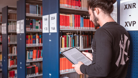 Foot von einem jungen Mann, der in einer Bibliothek steht und in der Hand ein Tablet hält.