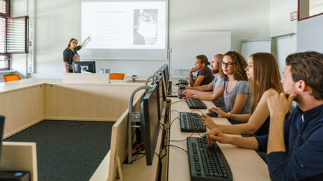 Foto von fünf Studierenden, die in einem Raum in einer Tischreihe an PCs sitzen. Eine Frau zeigt erklärend auf etwas an einer Beamer-Leinwand.__Five students are sitting in a room in a row of tables at PCs, a woman is pointing to something on a projector screen to explain.