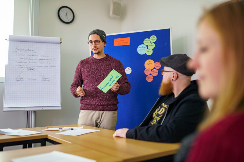 Foto eines Mitglieds der Arbeitsgruppe Ethnografie. Der junge Mann steht im Raum und hält einen grünen Zettel. Dahinter ein Flipchart und eine Pinnwand mit bunten angepinnten Zetteln. Rechts sitzen zwei Personen.