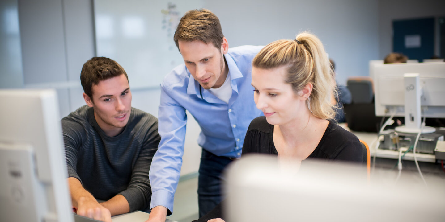 Photo of a professor standing between a male student and a female student in the computer room and explains something to both of them on the laptop. __ <br>The professor stands between a male student and a female student in the computer room and explains something to both of them on the laptop.